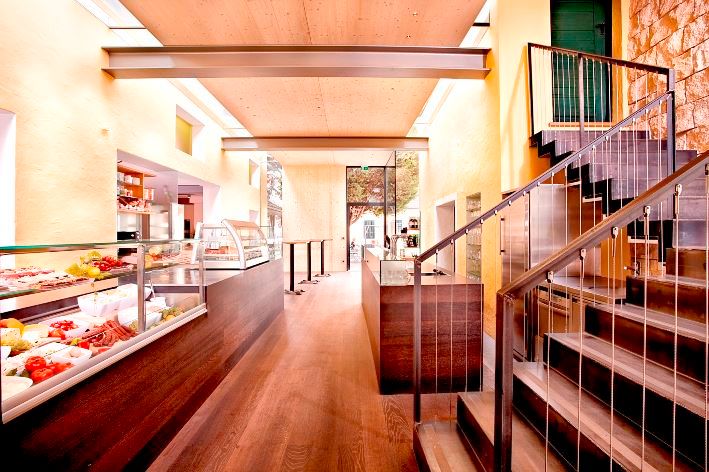 Pohled do interiéru moderního bufetu s dřevěnou podlahou, schody a skleněnými skříňkami.