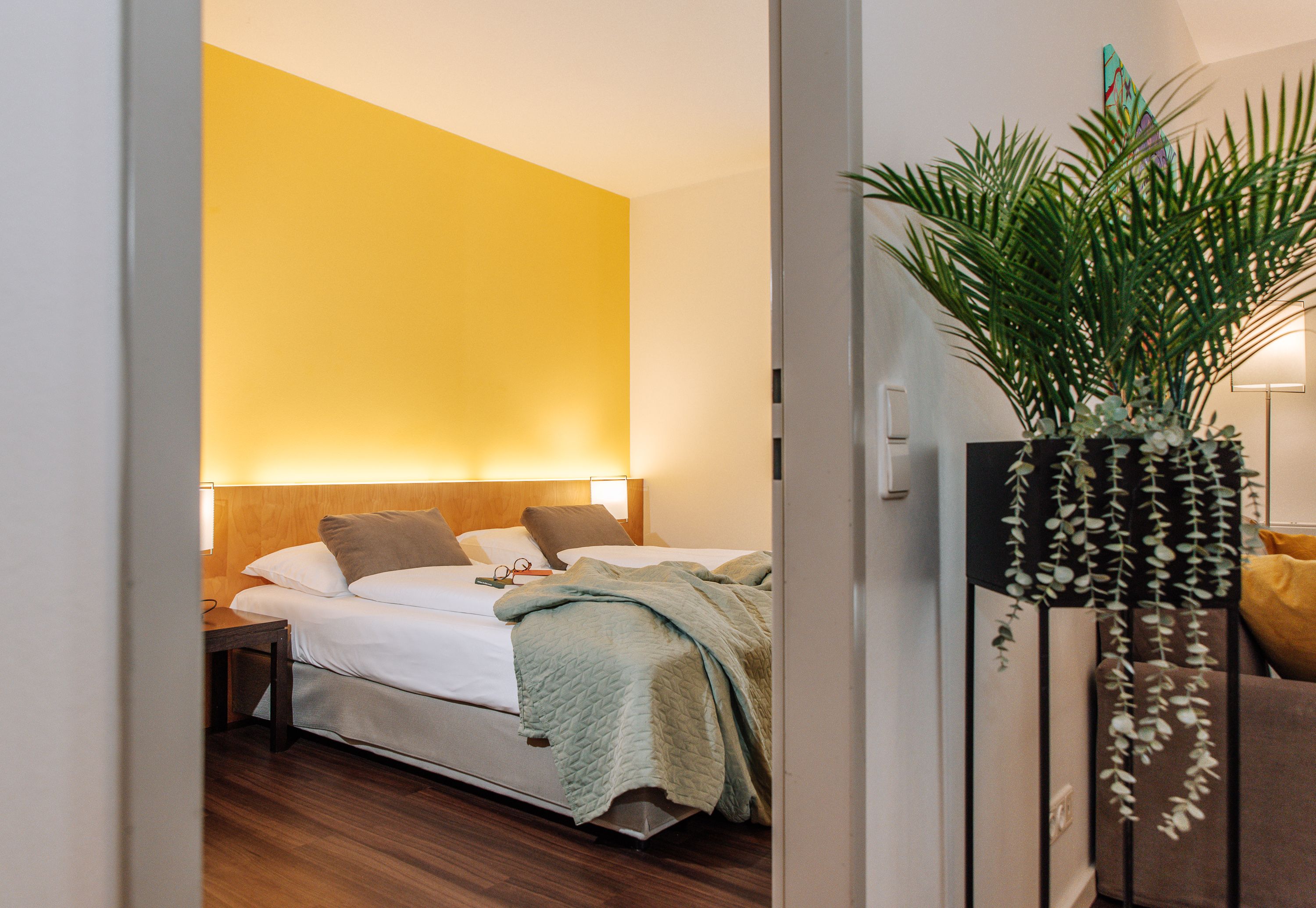 Hotelový pokoj se žlutou stěnou, manželskou postelí a rostlinou v popředí.