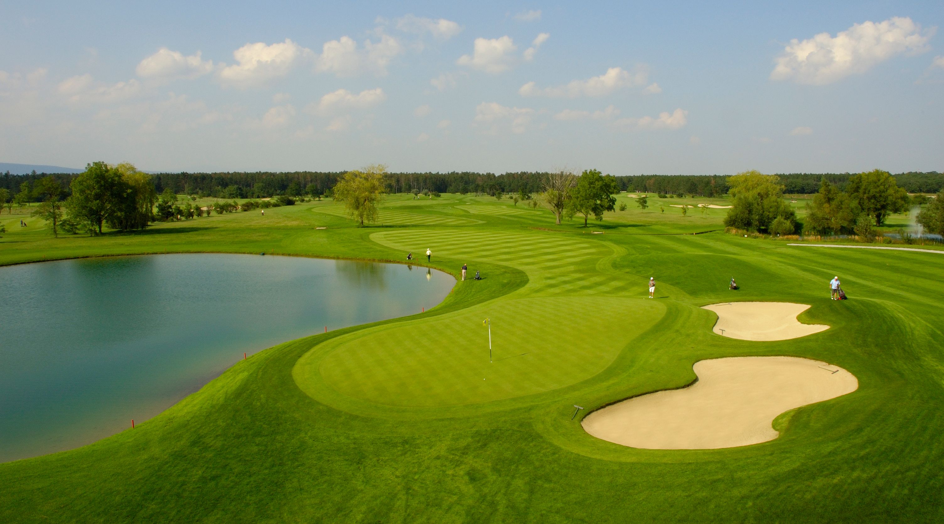 Golfové hřiště s rybníkem, písečnými bunkry a hráči.