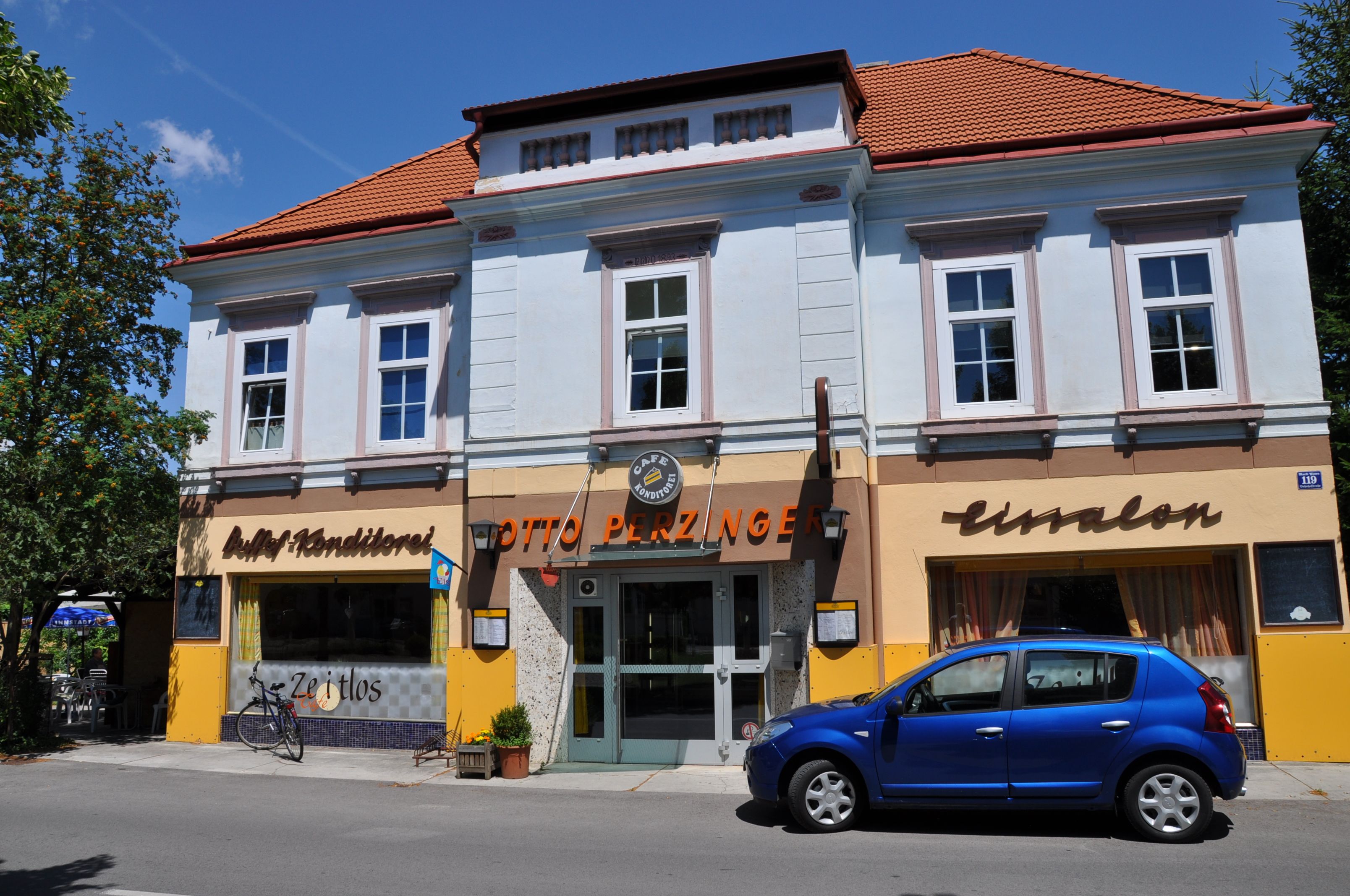Pohled zvenčí na kavárnu s modrou fasádou a červenou střechou, před níž parkuje modré auto.