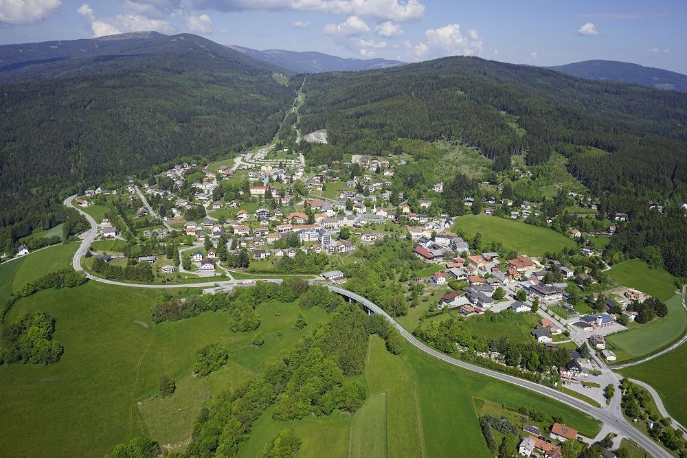 Letecký pohled na vesnici v zelené kopcovité krajině s lesy a loukami.