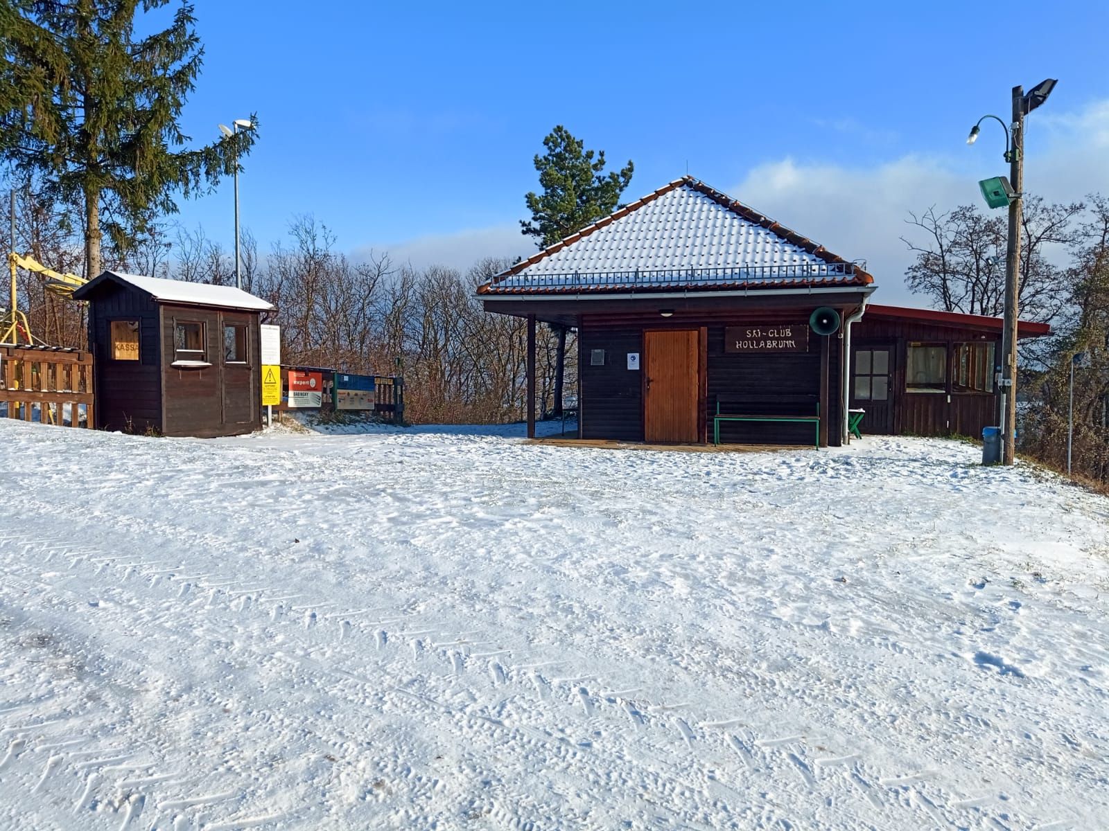 Zasněžená chata lyžařského klubu s dřevěnou chatou a stromy v pozadí.