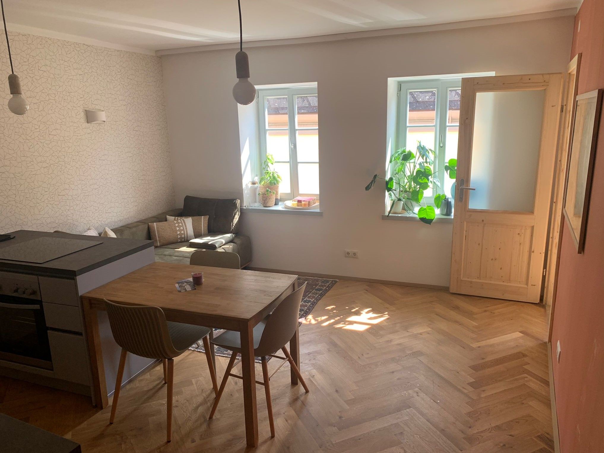 Světlý obývací pokoj s dřevěnou podlahou, jídelním stolem, pohovkou a rostlinami u okna.