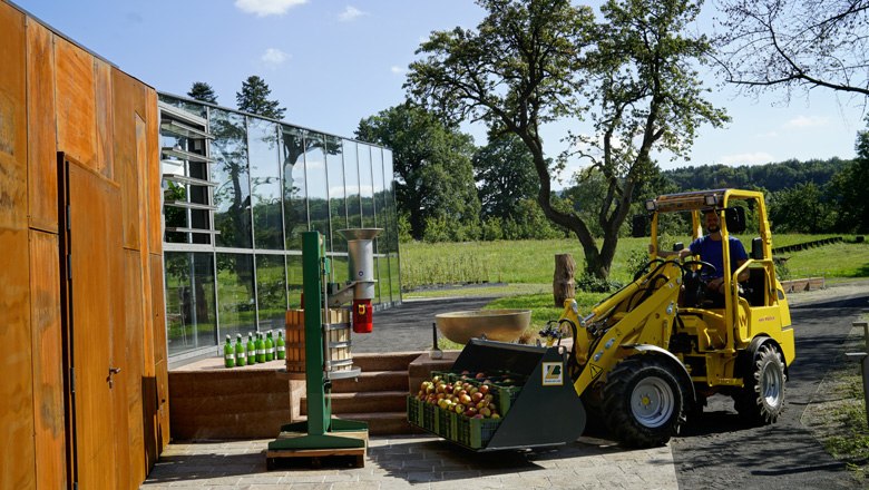 Žlutý elektrický Hoftrac převáží bedny s jablky před moderní budovou na farmě.