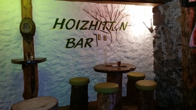 Rustik&aacute;ln&iacute; bar s dřevěn&yacute;m n&aacute;bytkem a n&aacute;pisem "Hoizhitt'n Bar" na zdi.