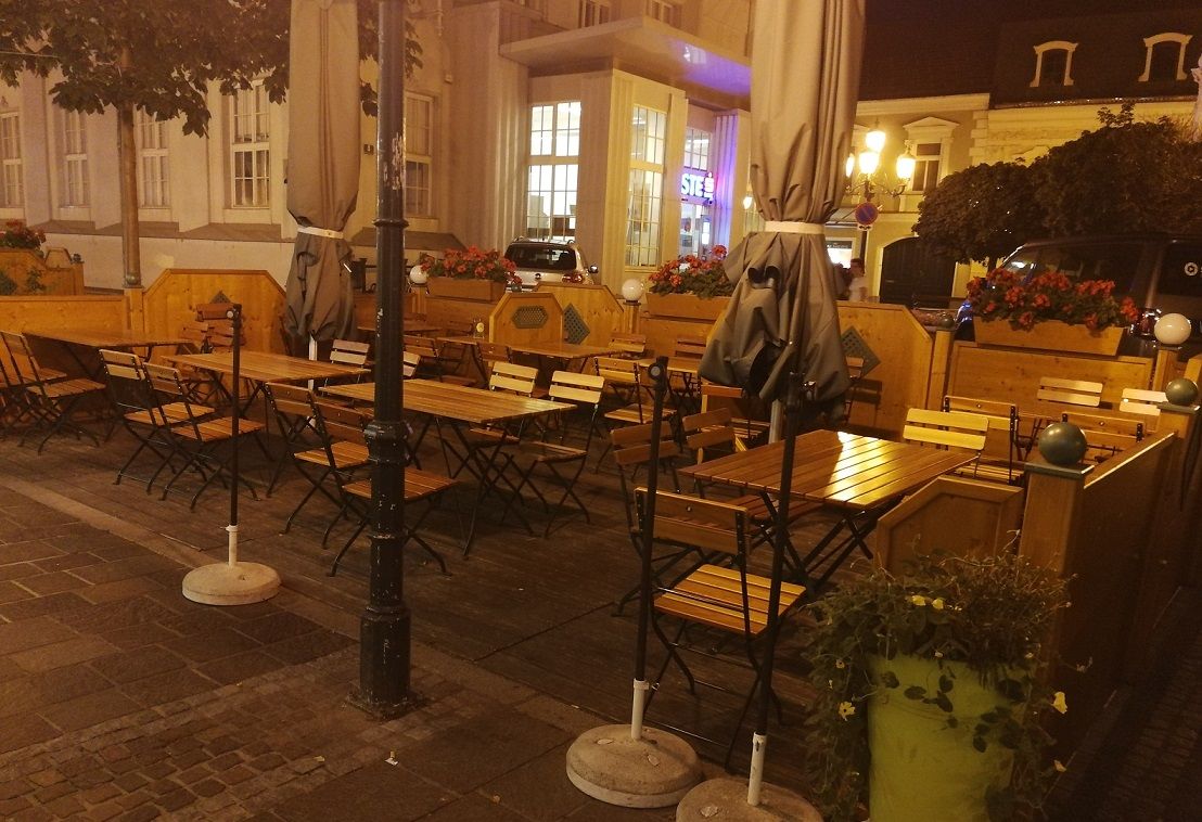 Venkovní prostor restaurace s prázdnými dřevěnými stoly a židlemi, obklopený rostlinami a nočními lucernami.