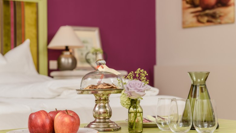 Stylově zař&iacute;zen&aacute; m&iacute;stnost se stolem, na kter&eacute;m jsou um&iacute;stěna jablka, su&scaron;enky pod skleněn&yacute;m zvonem, v&aacute;za s květinou a skleničky.