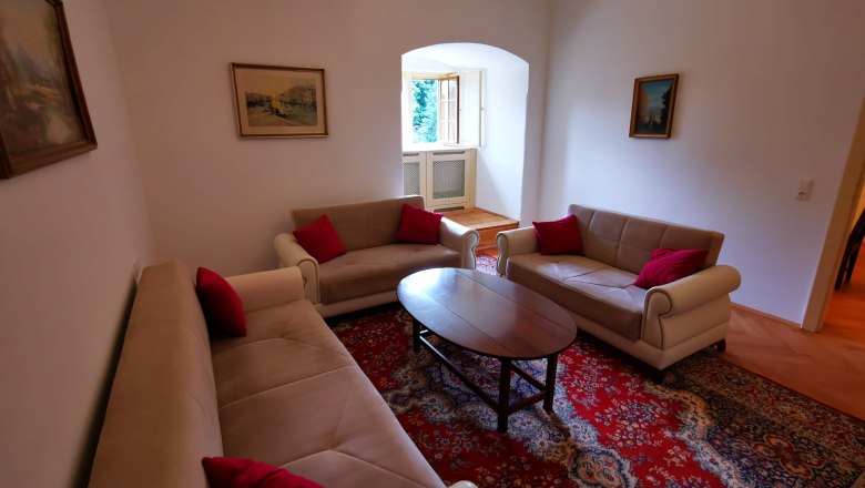 Ob&yacute;vac&iacute; pokoj se třemi pohovkami, červen&yacute;m kobercem a obrazy na stěn&aacute;ch.