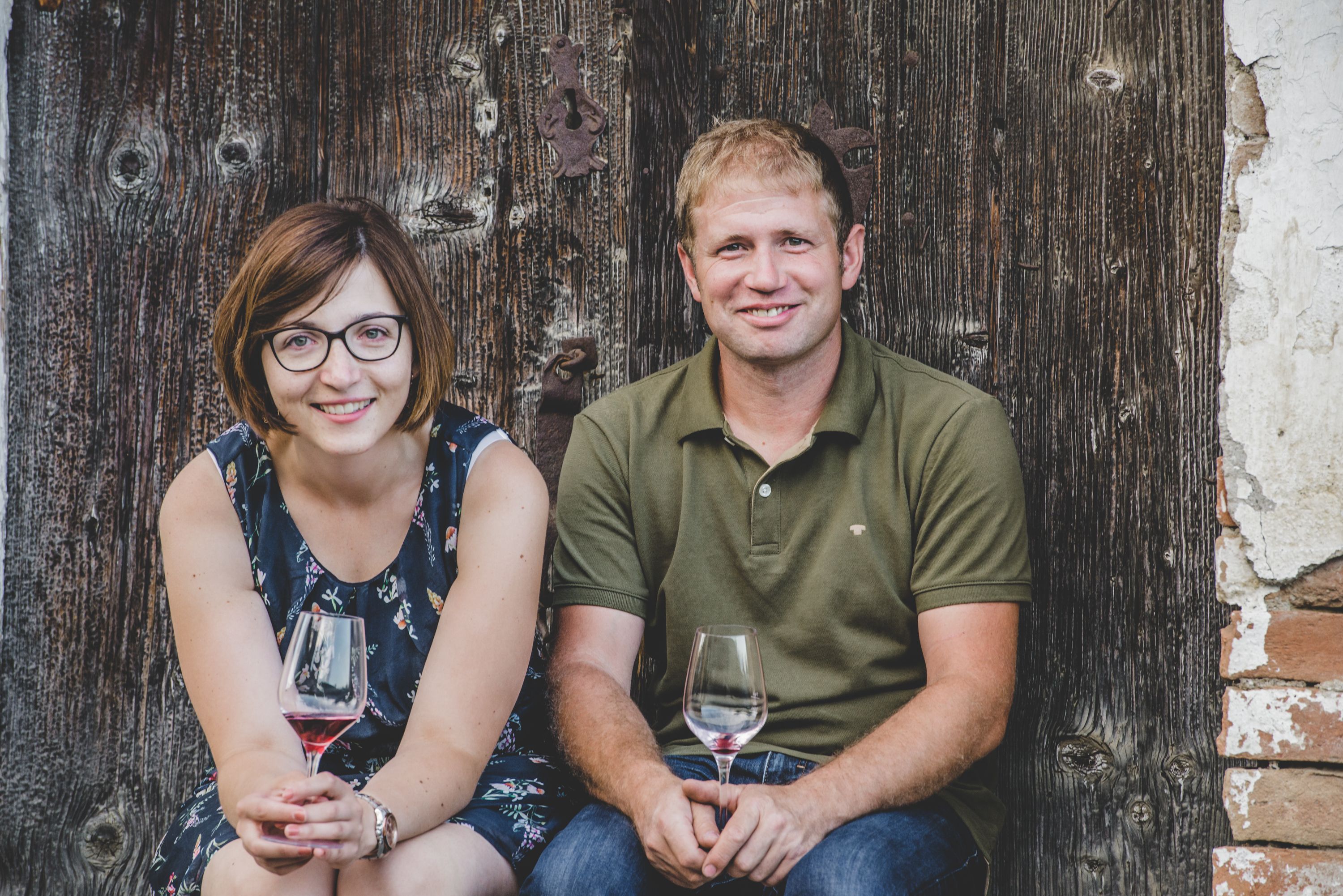 Muž a žena sedí před dřevěnou stěnou a v rukou drží skleničky na víno.