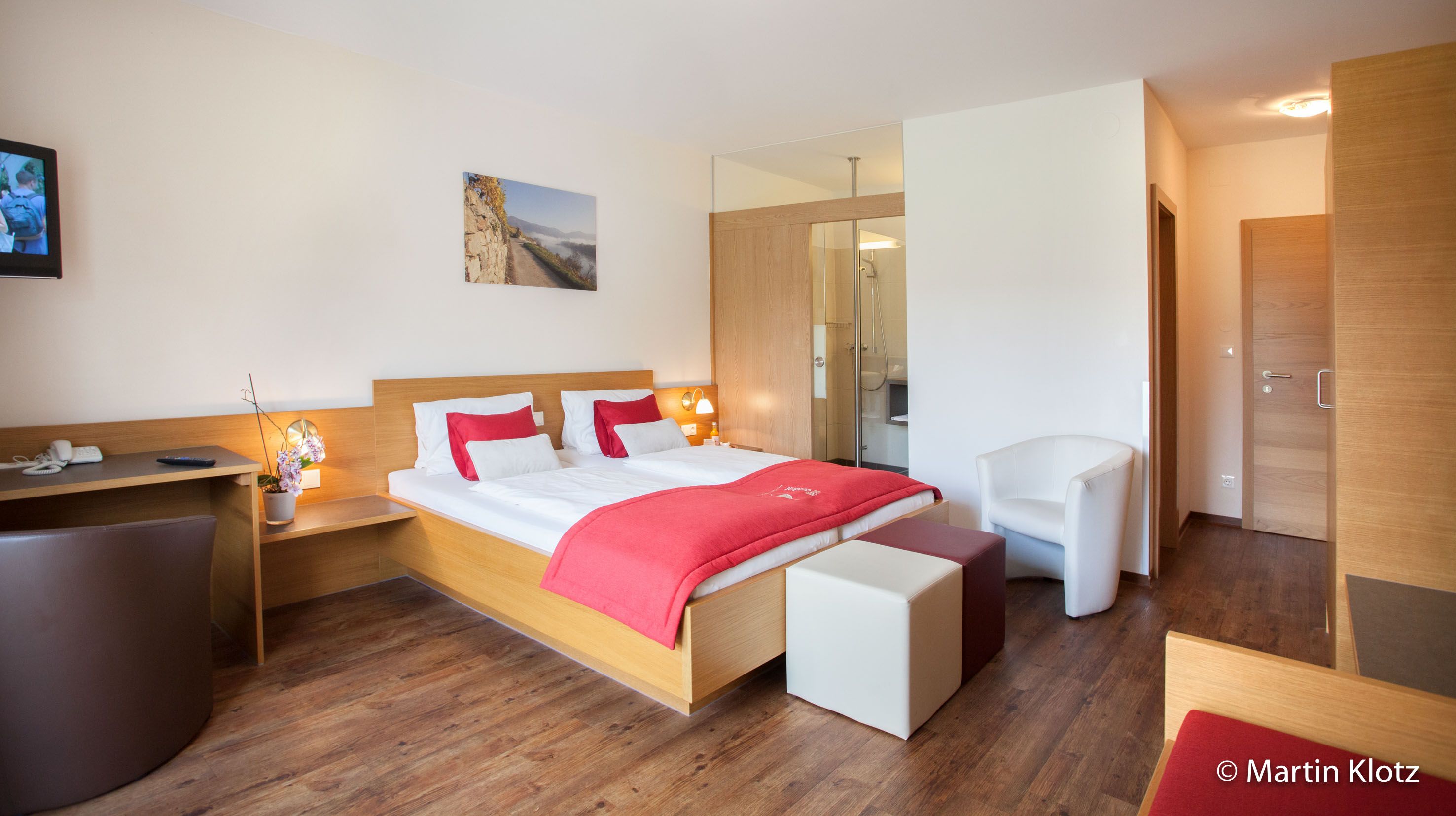 Moderní hotelový pokoj s manželskou postelí, červenými akcenty, psacím stolem a TV.