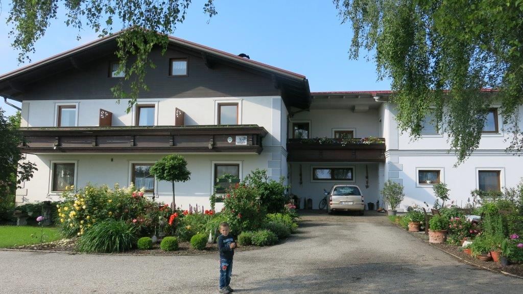 Tradiční penzion s balkonem, obklopený udržovanou zahradou. Na cestě před domem stojí dítě.