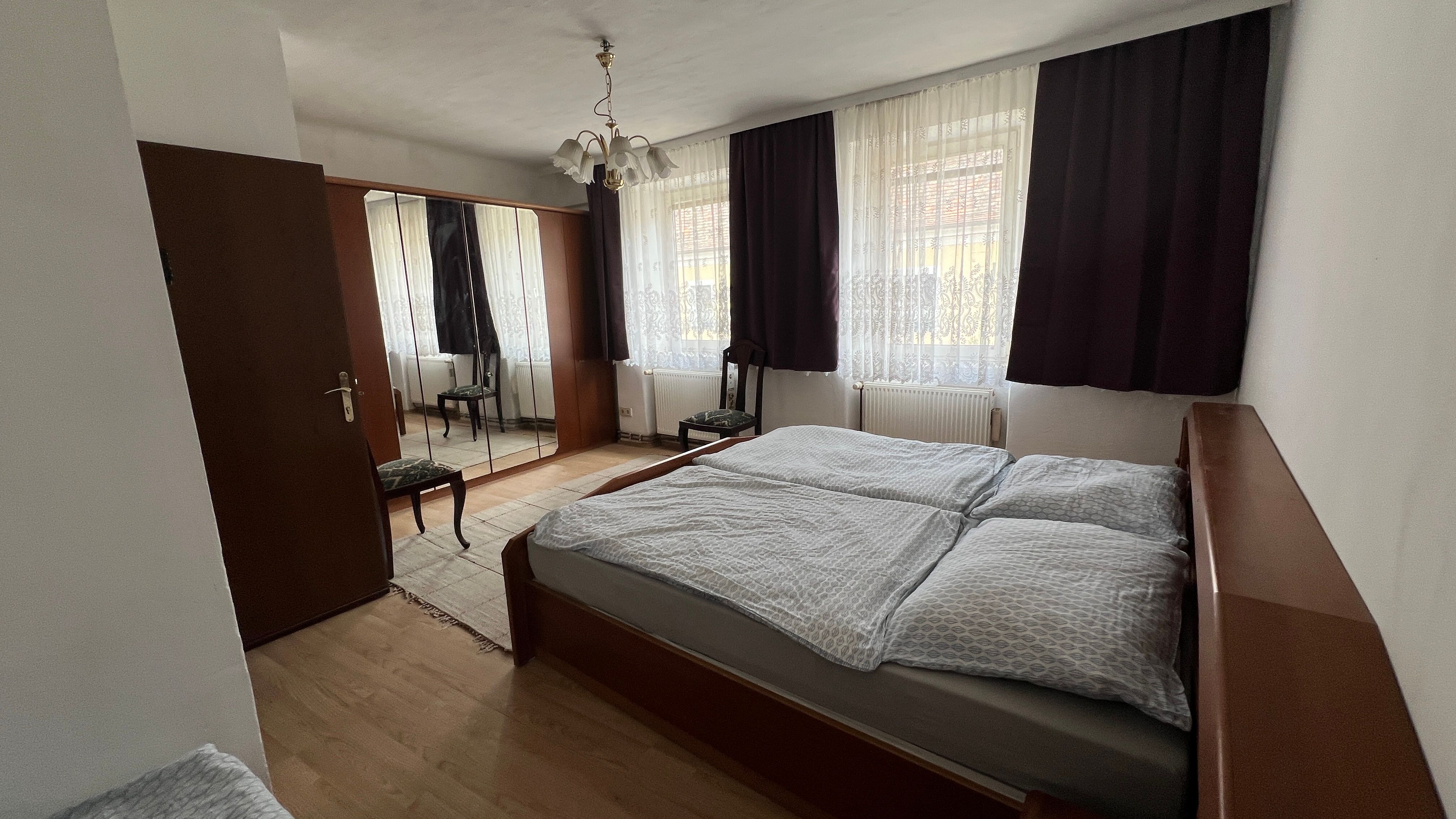 Ložnice s manželskou postelí, šatní skříní se zrcadlem, židlí a závěsy na oknech.