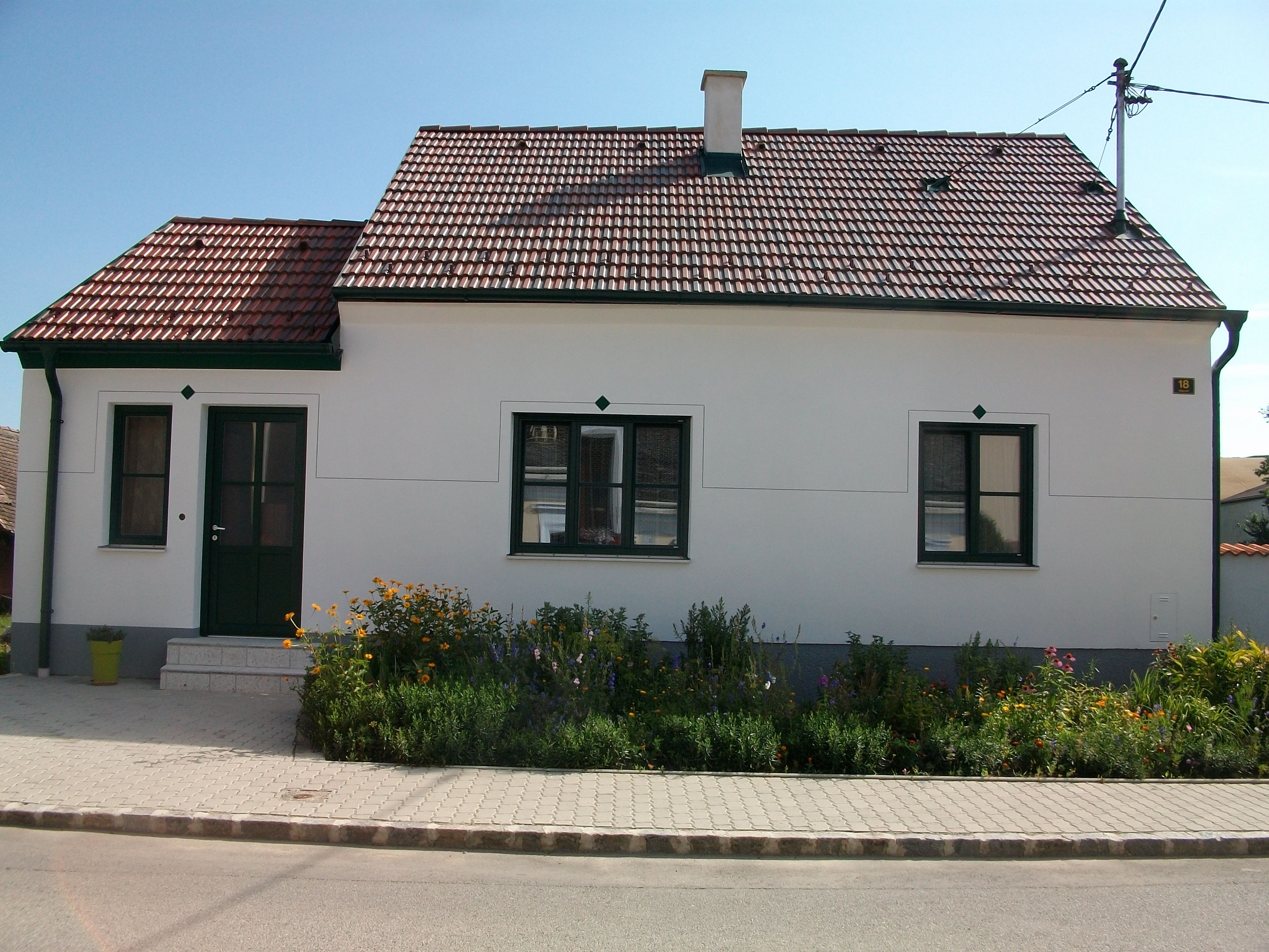 Malý bílý domek s červenou taškovou střechou a zeleným rámem dveří, obklopený udržovanou zahradou s květinami.