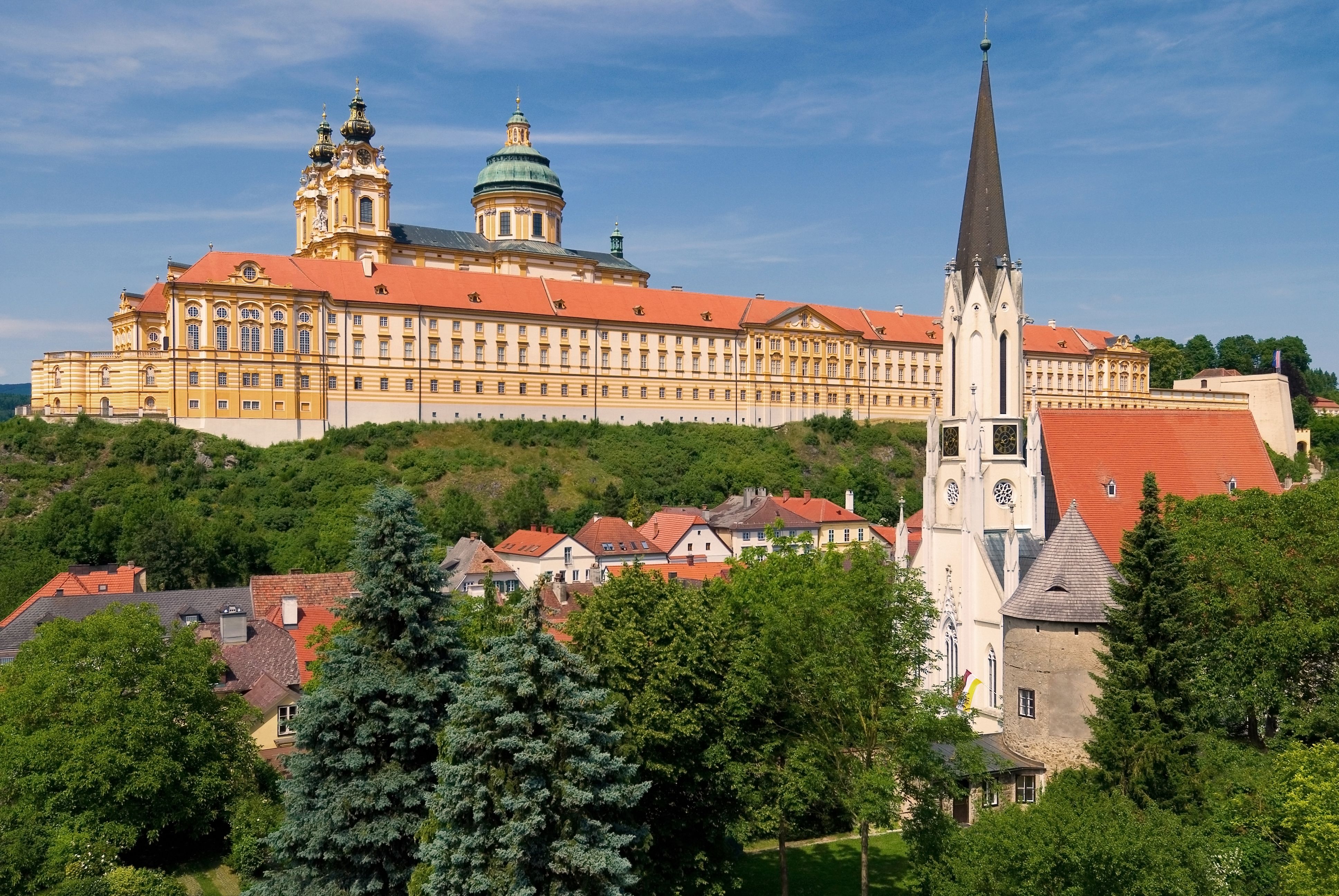 Pohled na opatství Melk a farní kostel v Melku, obklopené zelenou krajinou a modrou oblohou.