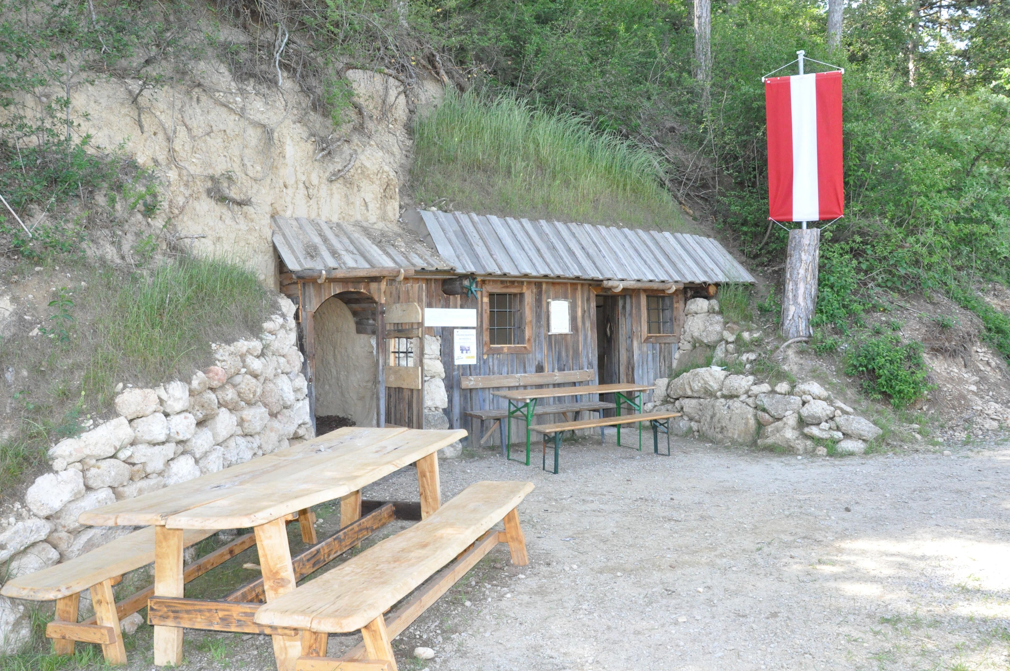 Rustikální dřevěná chata se stolem a lavicemi před ní, postavená do kopce. Na stromě visí červenobílá vlajka.