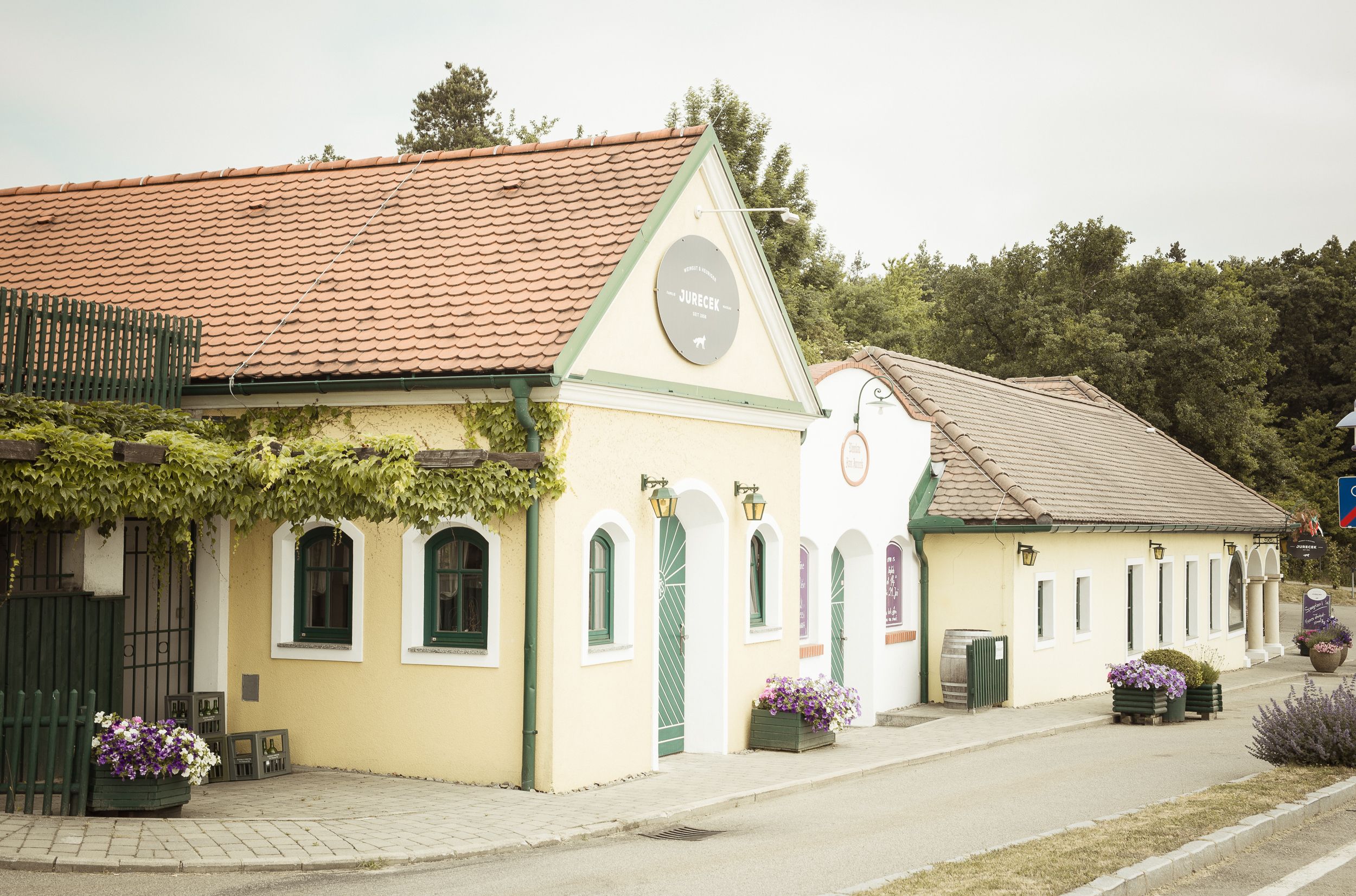Pohled zvenčí na tradiční heuriger se žlutou fasádou a červenými střešními taškami, obklopenou rostlinami.