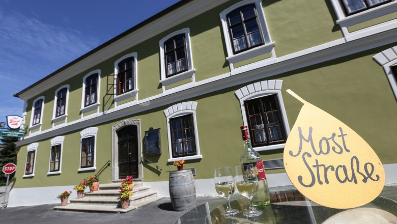 Zelen&aacute; budova s b&iacute;l&yacute;mi okny, před n&iacute; stůl s lahv&iacute; v&iacute;na a sklenicemi, cedule s n&aacute;pisem "Most Stra&szlig;e".