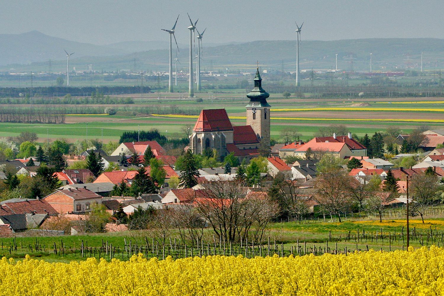 Pohled na Pillichsdorf s kostelem, obklopený poli a větrnými turbínami v pozadí.