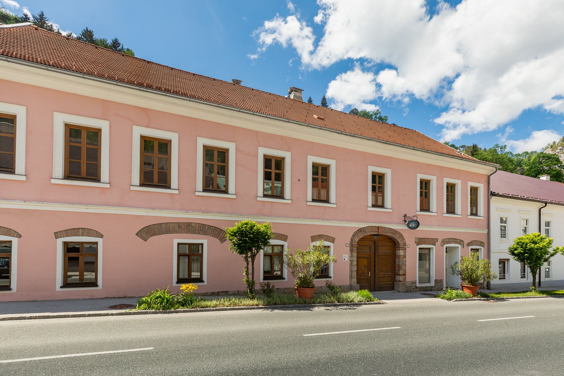 Dvoupatrová růžová budova s hnědými okny a dřevěnými dveřmi, obklopená rostlinami, pod modrou oblohou.