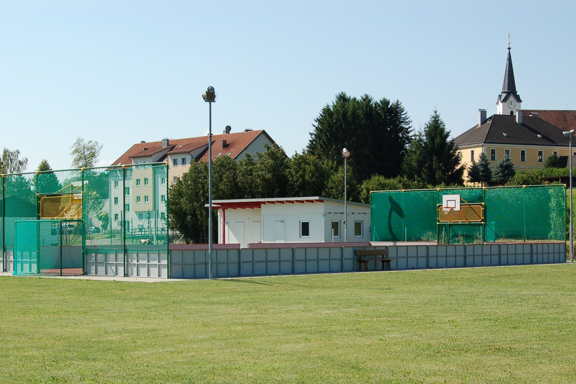 Zábavné hřiště s basketbalovým košem a zelenou sítí vedle malé budovy. V pozadí jsou vidět obytné budovy a kostel.