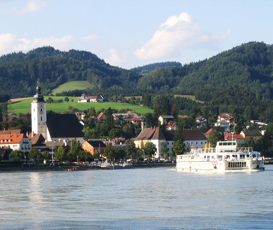 Město Grein s kostelem a lodí na Dunaji, obklopené kopci.