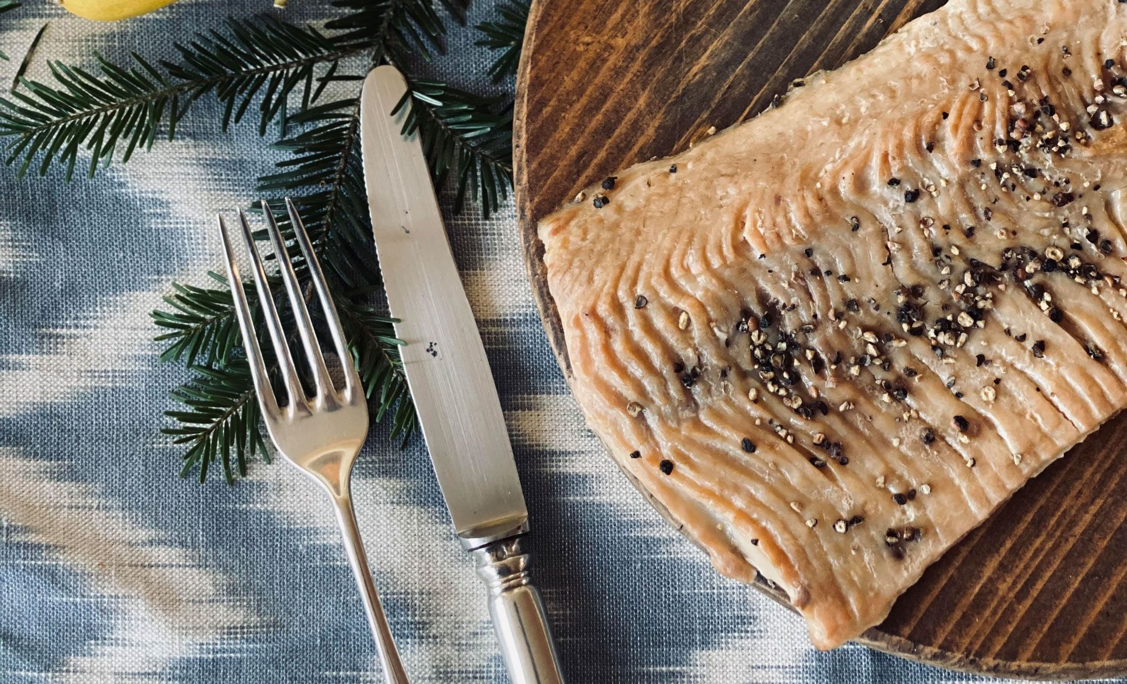 Detailní záběr na uzený filet z kapra na dřevěném talíři, zdobený zrnky pepře. Vedle něj jsou na modré utěrce položeny vidlička a nůž.