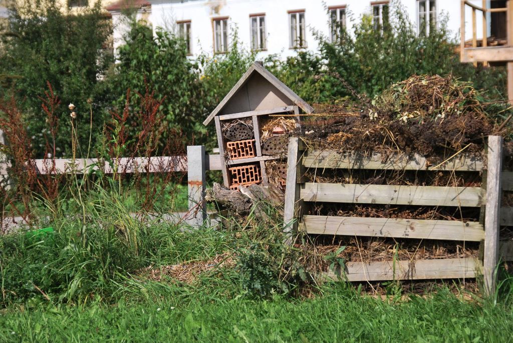 Hmyzí hotel vedle kompostu na zahradě se zelenou trávou a stromy v pozadí.