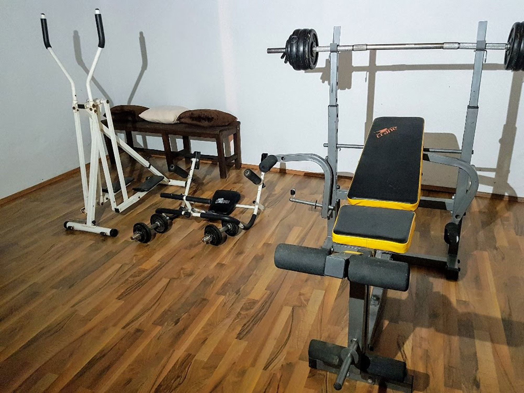Posilovna s posilovací lavicí, crossovým trenažérem a činkami na dřevěné podlaze.