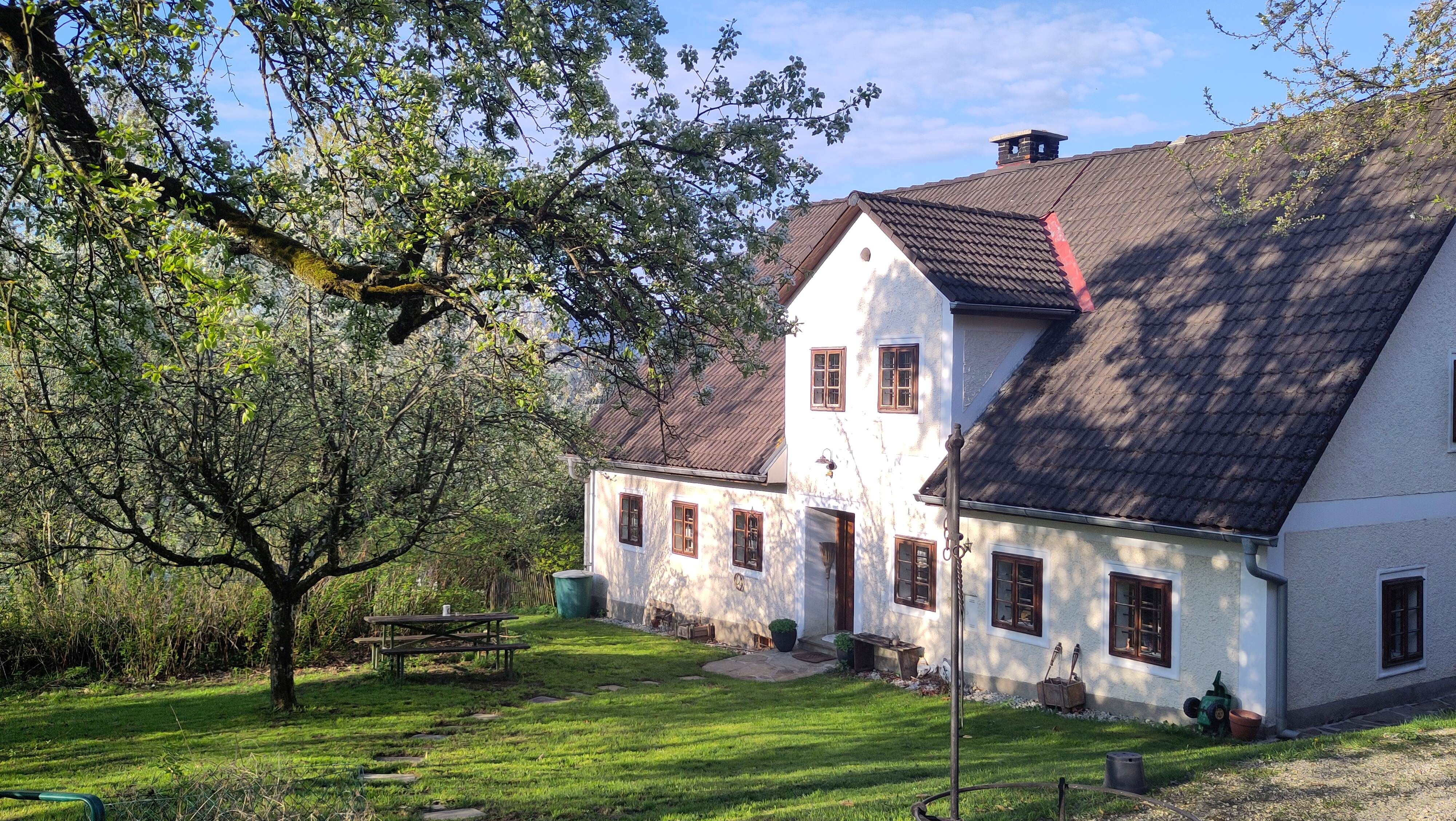 Tradiční venkovský dům s bílými stěnami a hnědými okny, obklopený zelenými stromy a upravenou zahradou s piknikovým stolem.