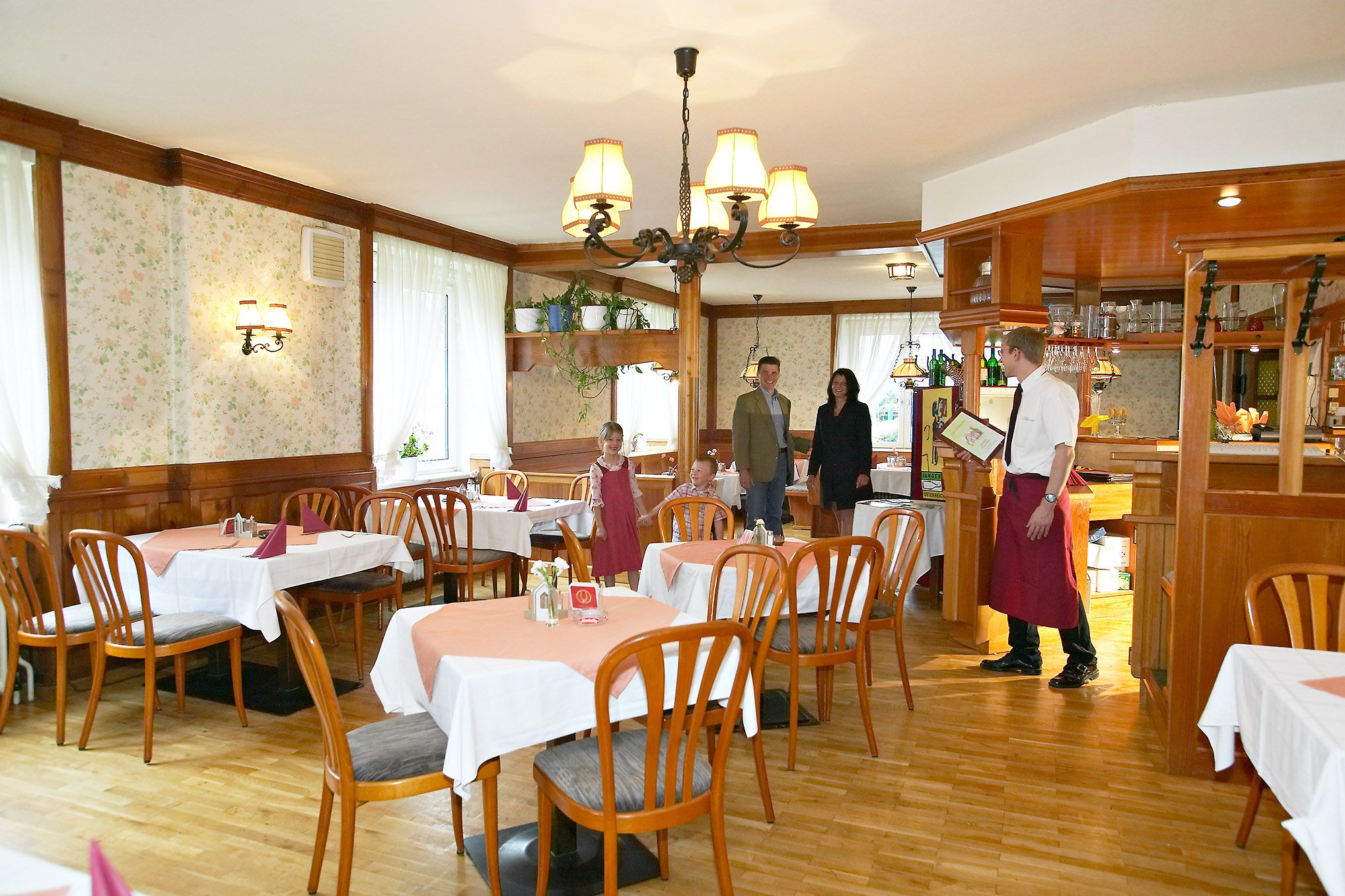 Rustikální restaurace s dřevěným nábytkem a ubrusy. Číšník vítá rodinu u vchodu.