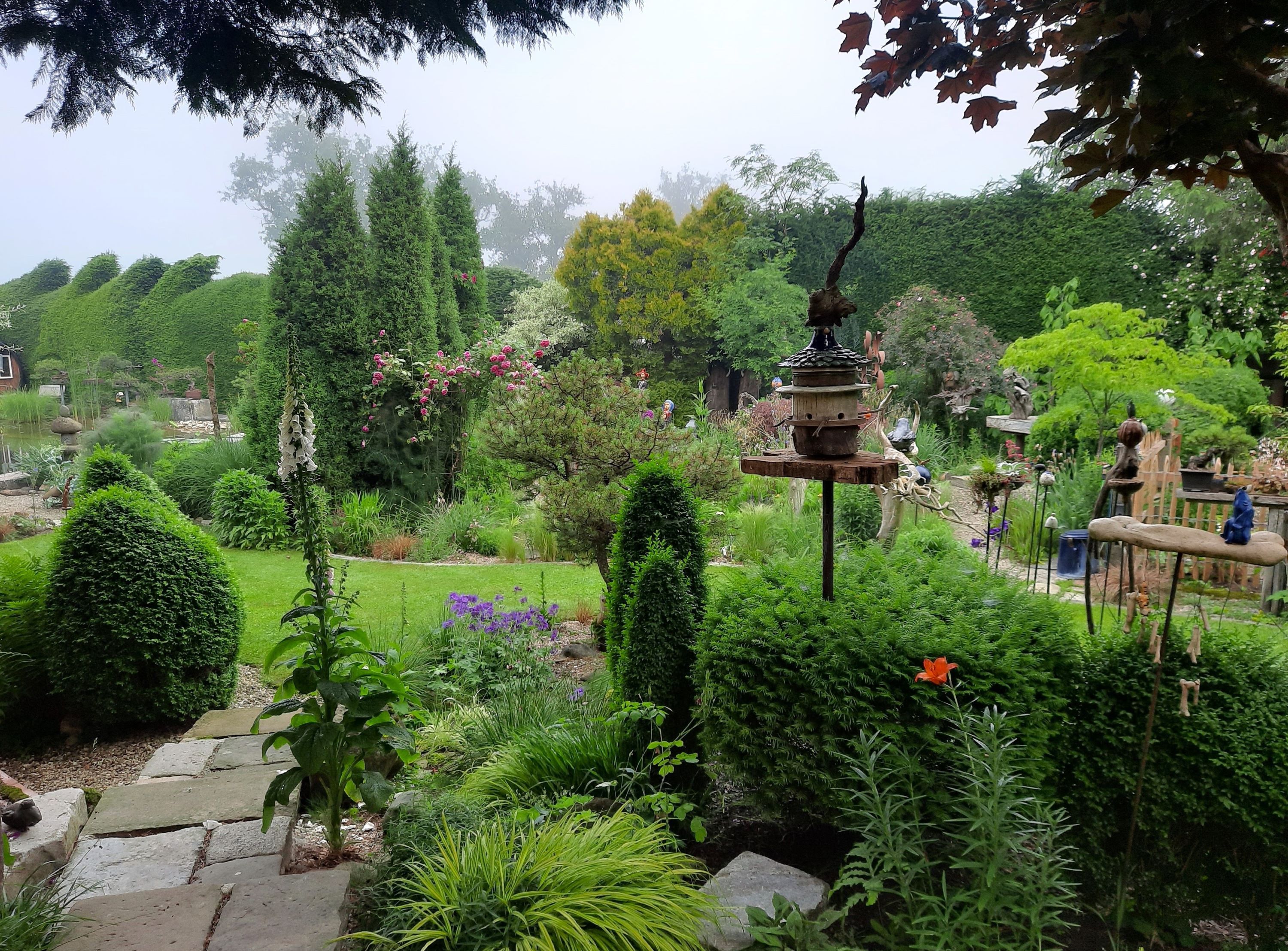 Svěží zahrada s různými rostlinami, stromy a dekoracemi. V centru pozornosti je ptačí budka.