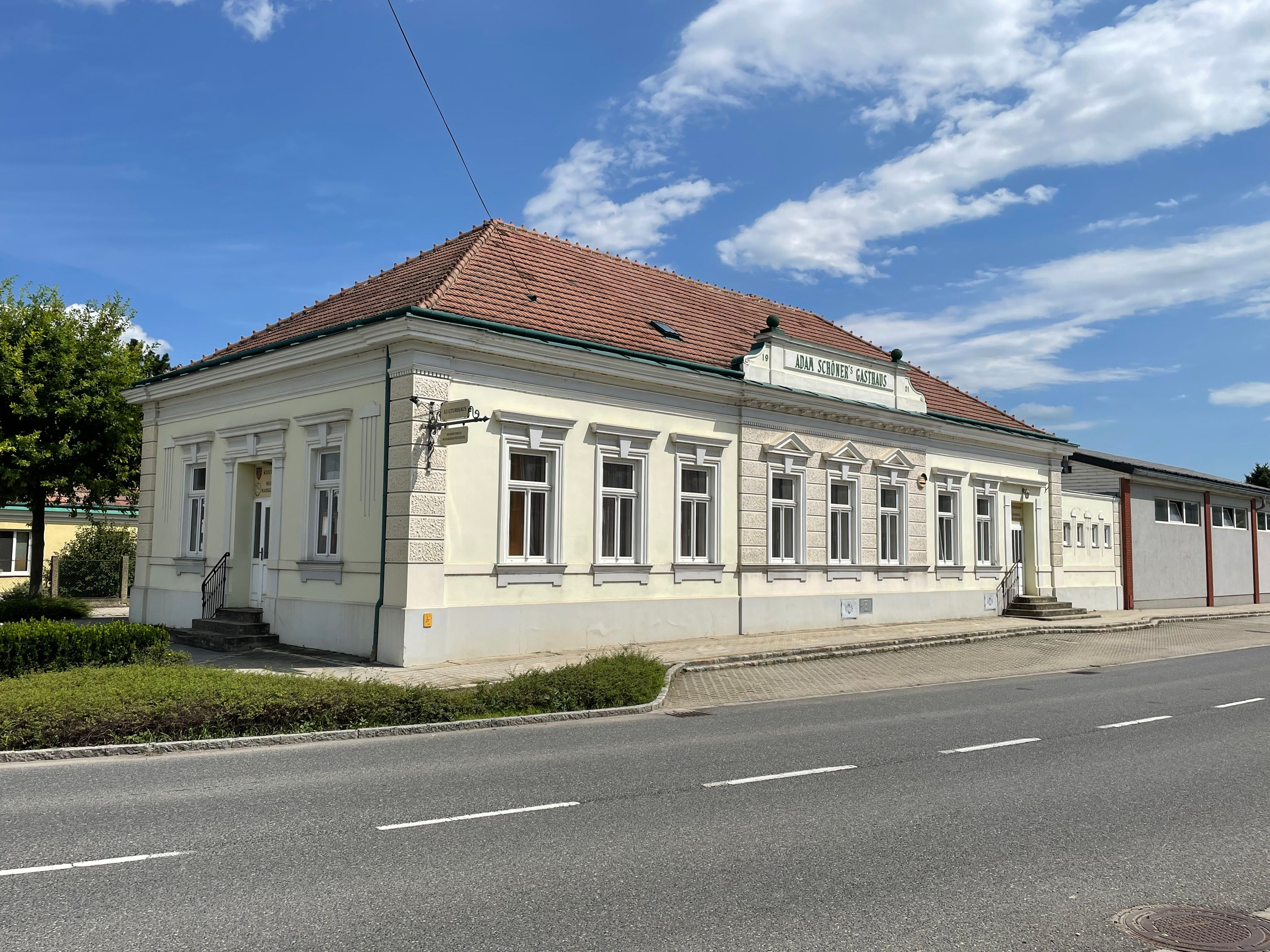 Kulturní dům, světlá historická budova s červenou střechou z pálených tašek, stojí u silnice pod modrou oblohou.