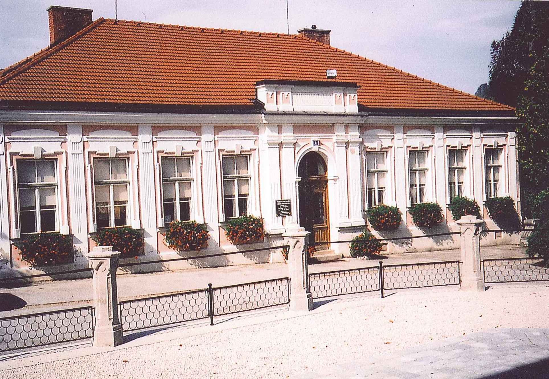 Historická budova s červenou taškovou střechou a bílými okenními rámy, obklopená květinami.