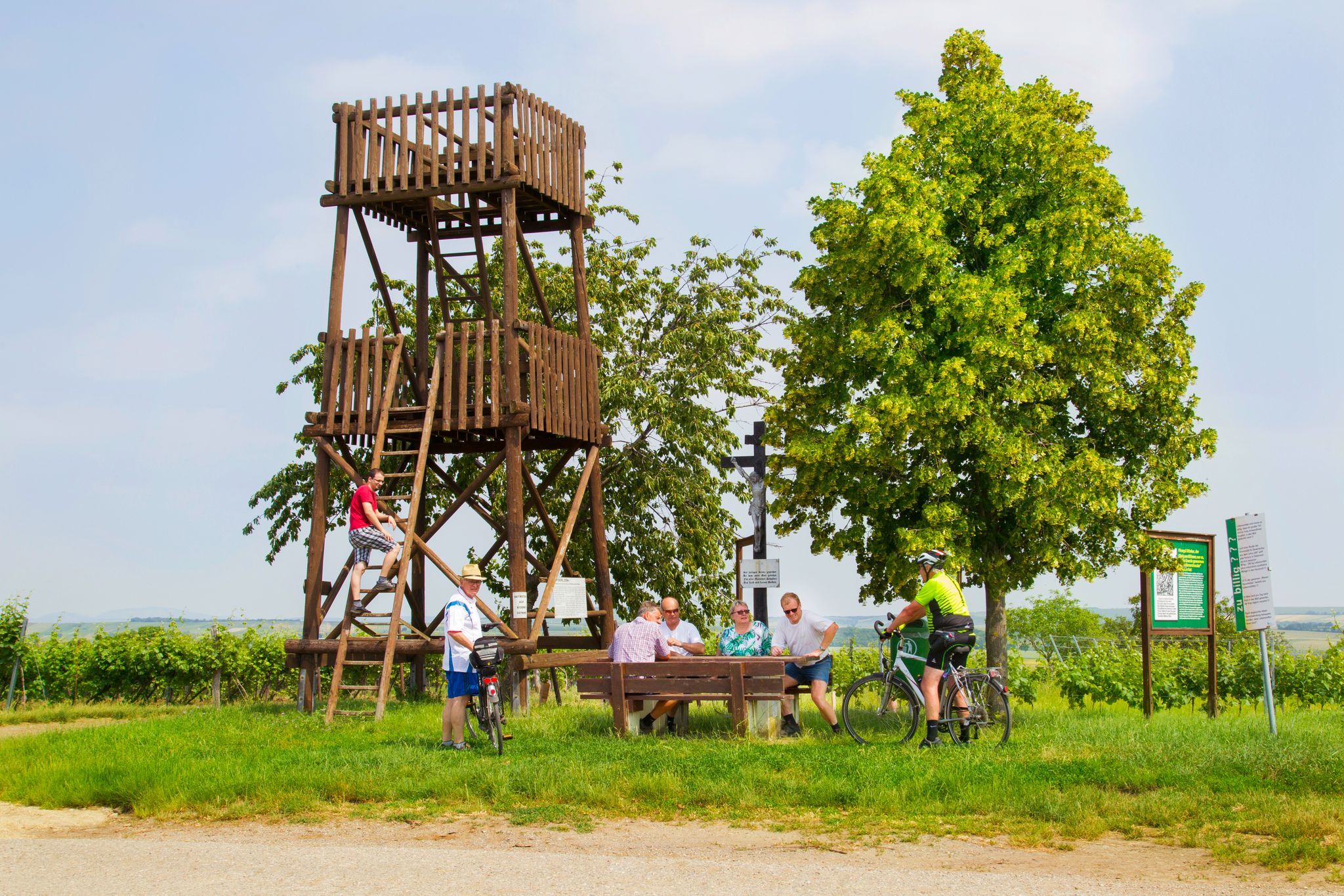 Lidé odpočívají u dřevěné věže a stolu venku, obklopeni stromy a cedulemi.
