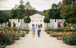 Zwischen blühenden Rosen im Doblhoffpark Baden spazieren eine Frau und ein Mann, vertieft in ein Gespräch. Im Hintergrund erhebt sich die eindrucksvolle Fotoausstellung von La Gacilly Baden Photo Baden, deren große Bilder das Szenario bereichern.