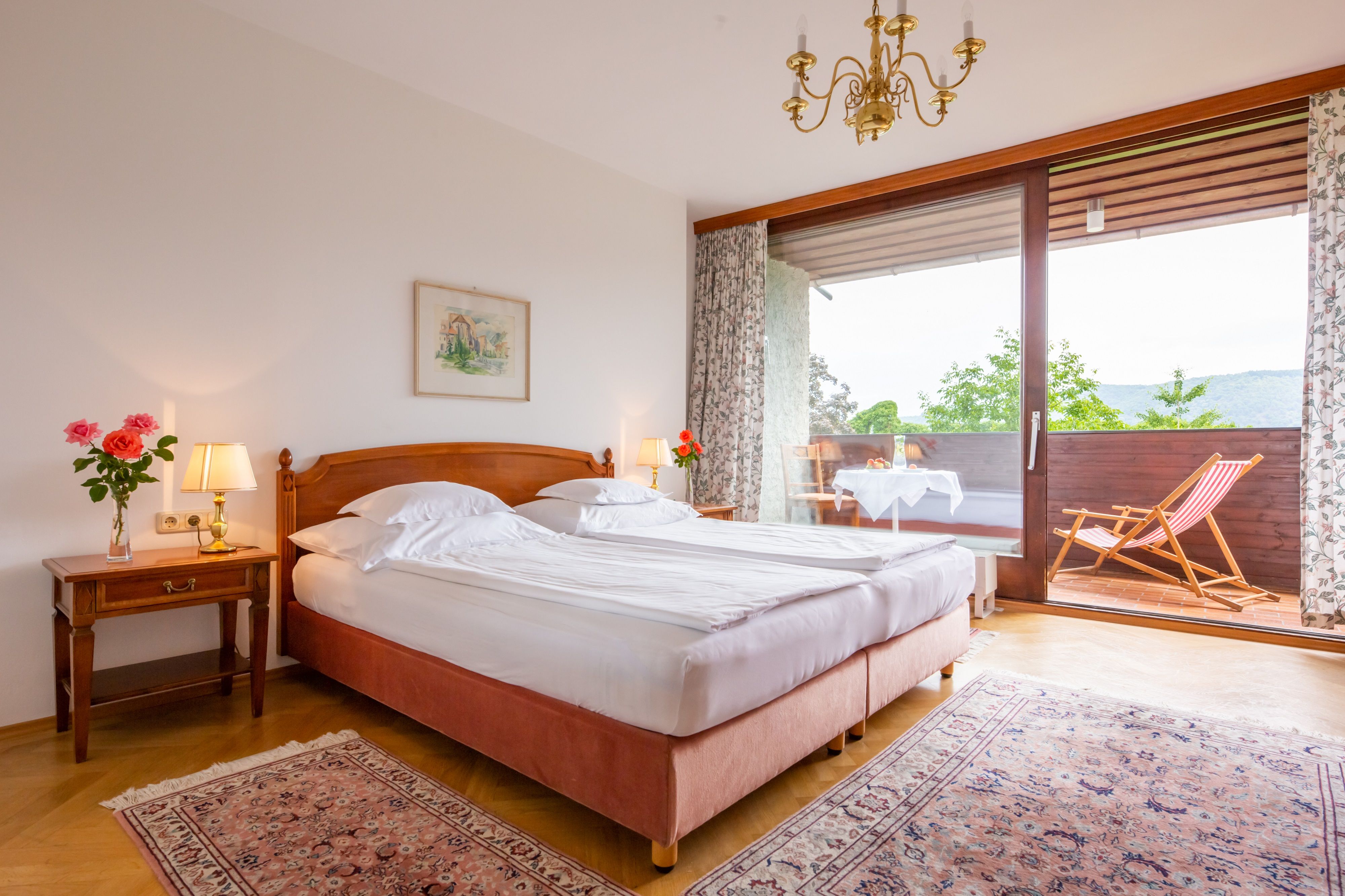 Útulná ložnice s manželskou postelí, nočními lampičkami a balkonem s lehátkem a stolem.