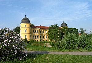 Žlutý hrad se dvěma věžemi a červenou střechou, obklopený stromy a květinami.
