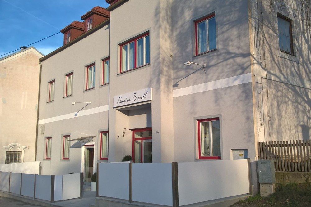 Vnější pohled na penzion Brandl s šedou fasádou a červenými okenními rámy.