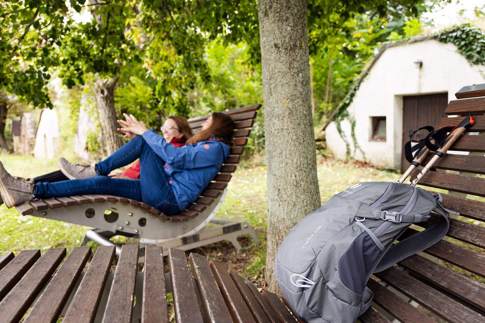 Dvě ženy na dřevěném lehátku, turistický batoh a staré vinné sklepy v pozadí. 