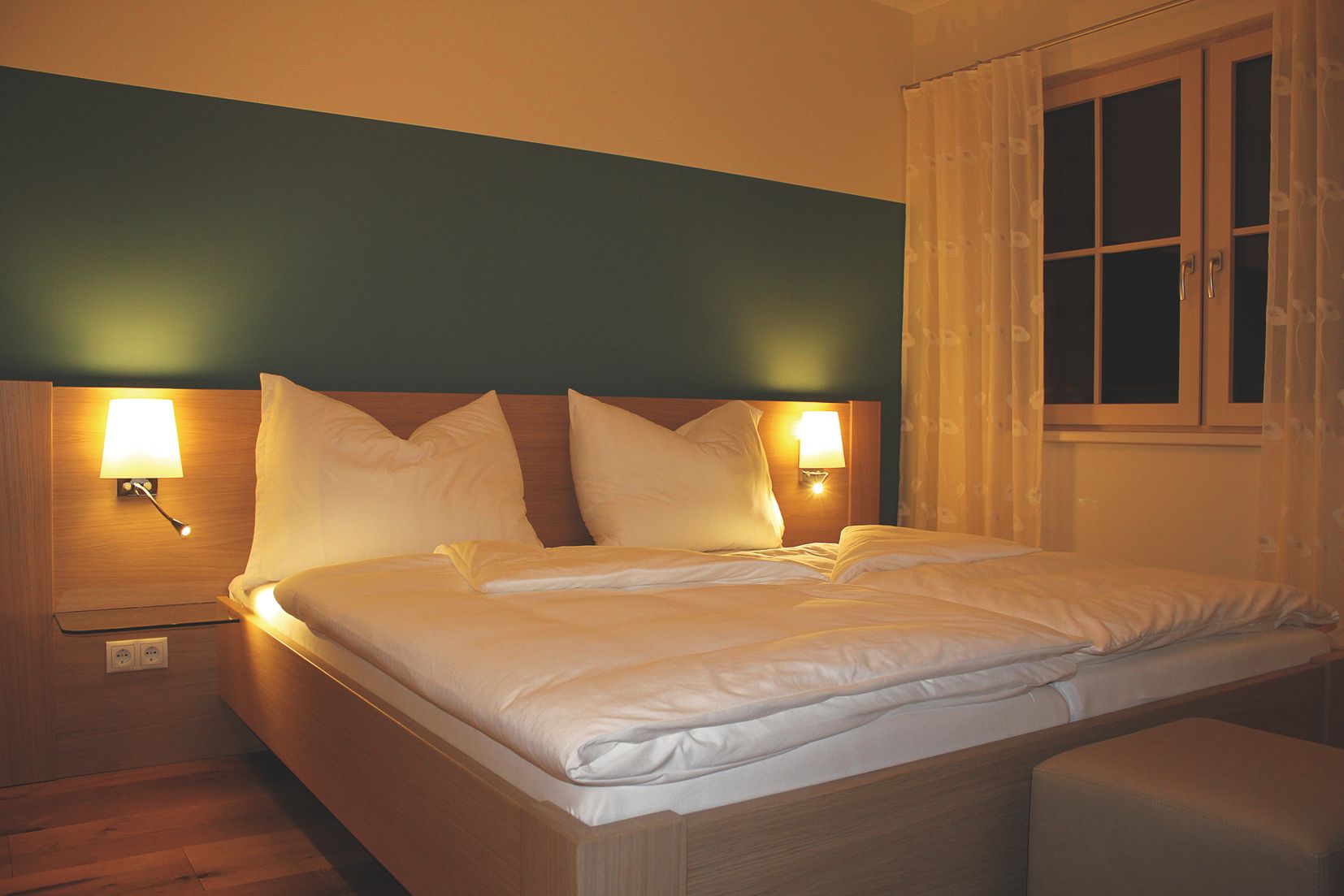 Útulný pokoj pro hosty s manželskou postelí, dvěma nočními lampičkami a oknem se závěsy.