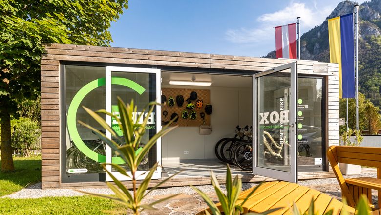 Moderní box na kola s otevřenými dveřmi, koly a přilbami, obklopený přírodou a vlajkami.