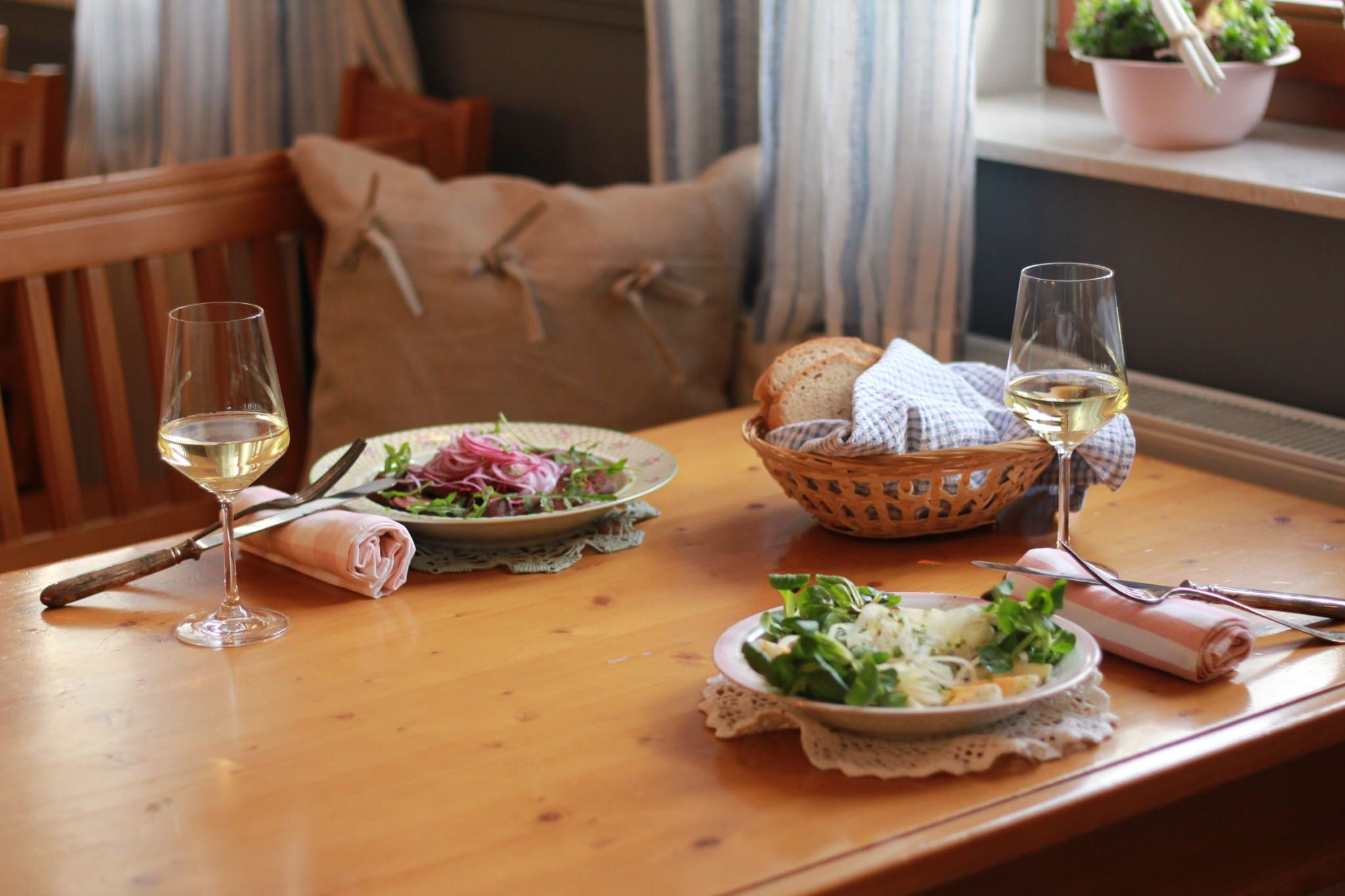 Dva talíře salátu a dvě sklenice bílého vína na dřevěném stole, vedle nich košík s chlebem.