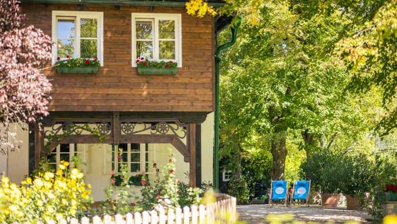 Idylický venkovský dům s dřevěnými prvky, obklopený kvetoucími zahradami a stromy. Na zahradě jsou dvě modrá lehátka.