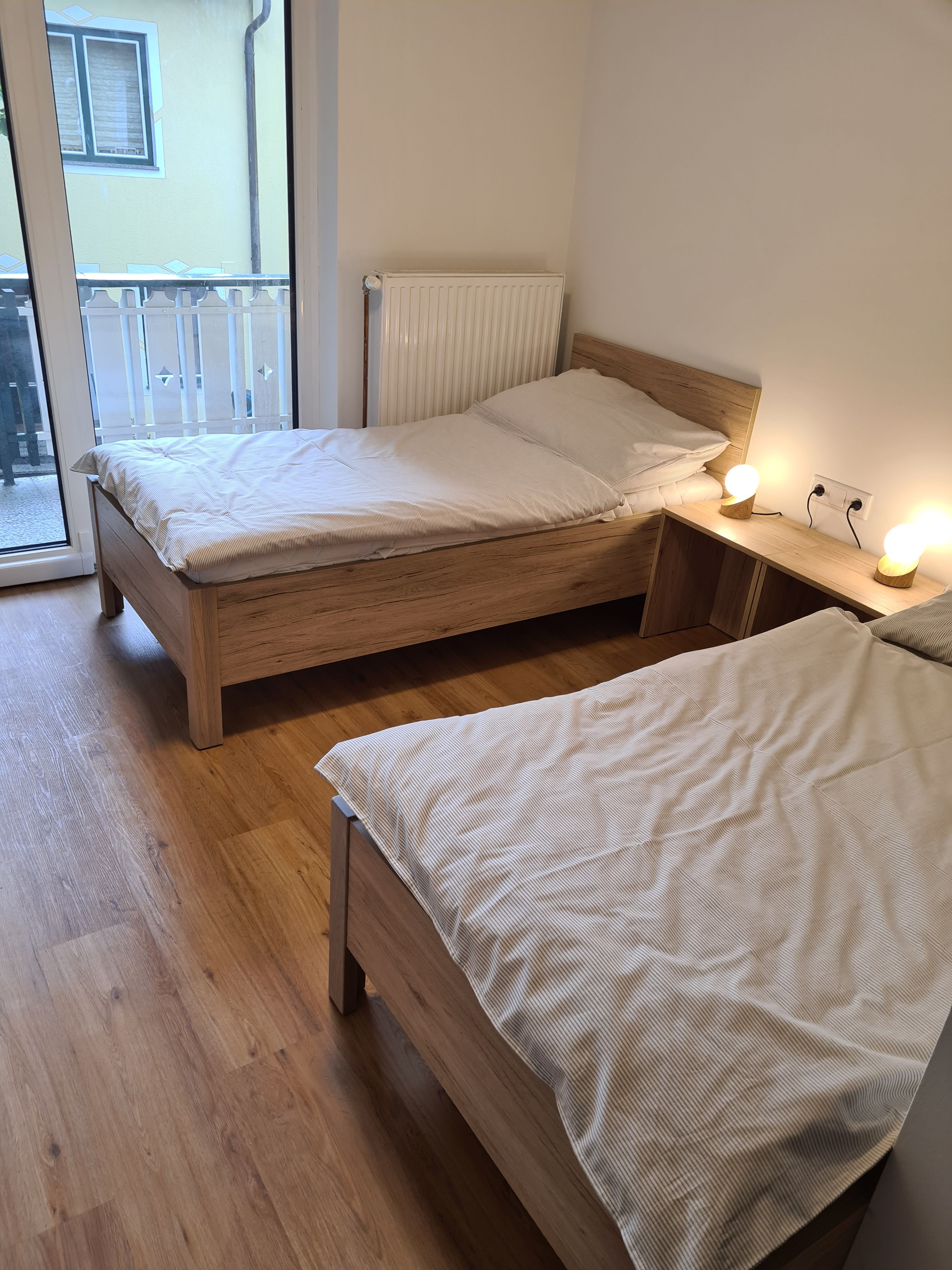 Dvě samostatné postele ve světlém pokoji s dřevěnou podlahou a balkonem.