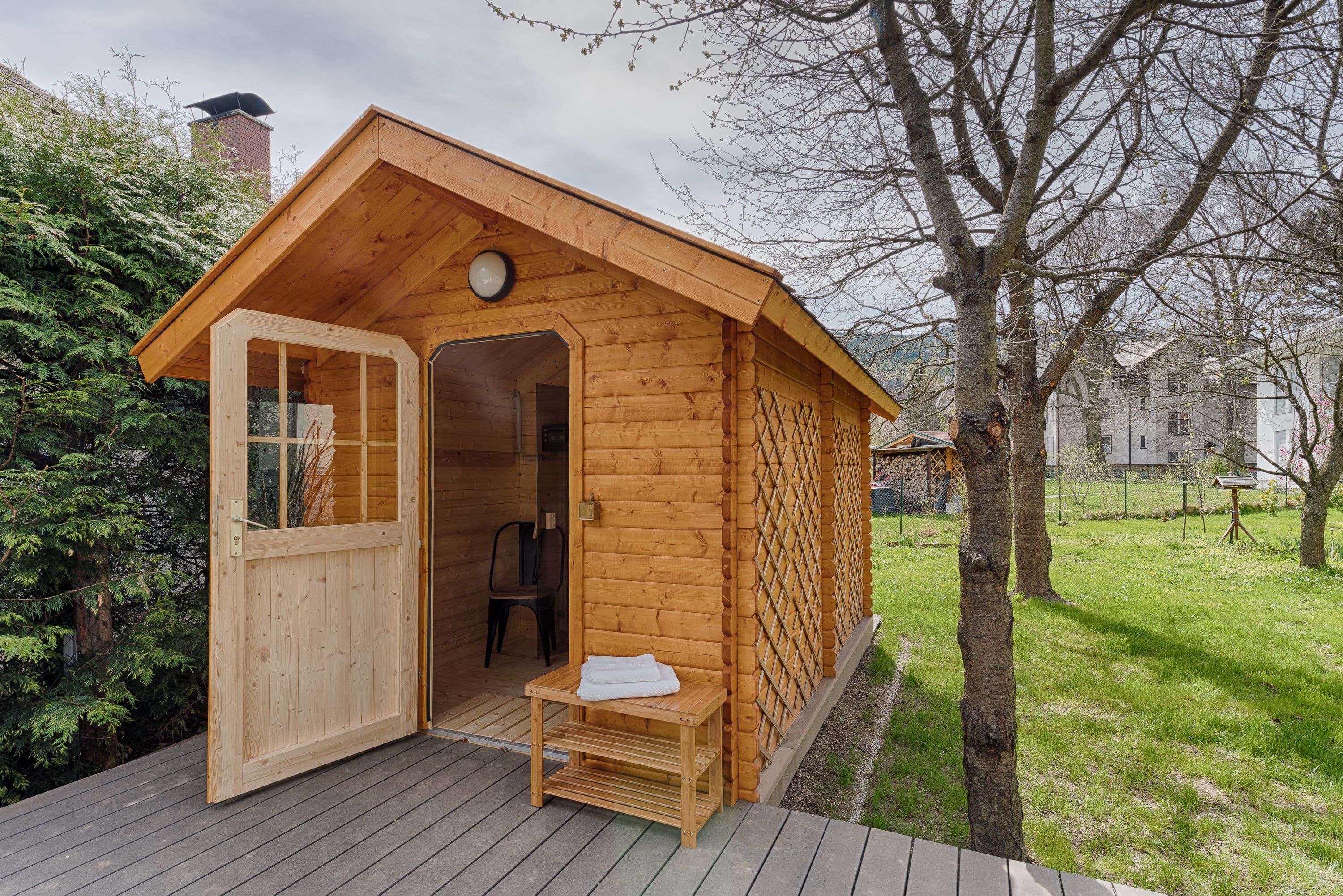 Malá dřevěná sauna v zahradě s otevřenými dveřmi a ručníky na lavičce před ní.