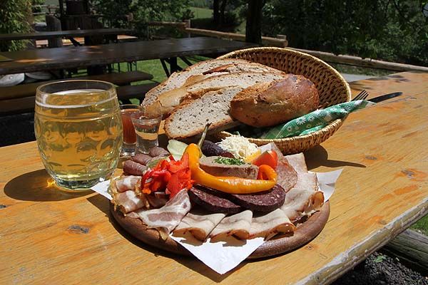 Občerstvení s chlebem, klobásou, slaninou a nápoji na venkovním dřevěném stole.