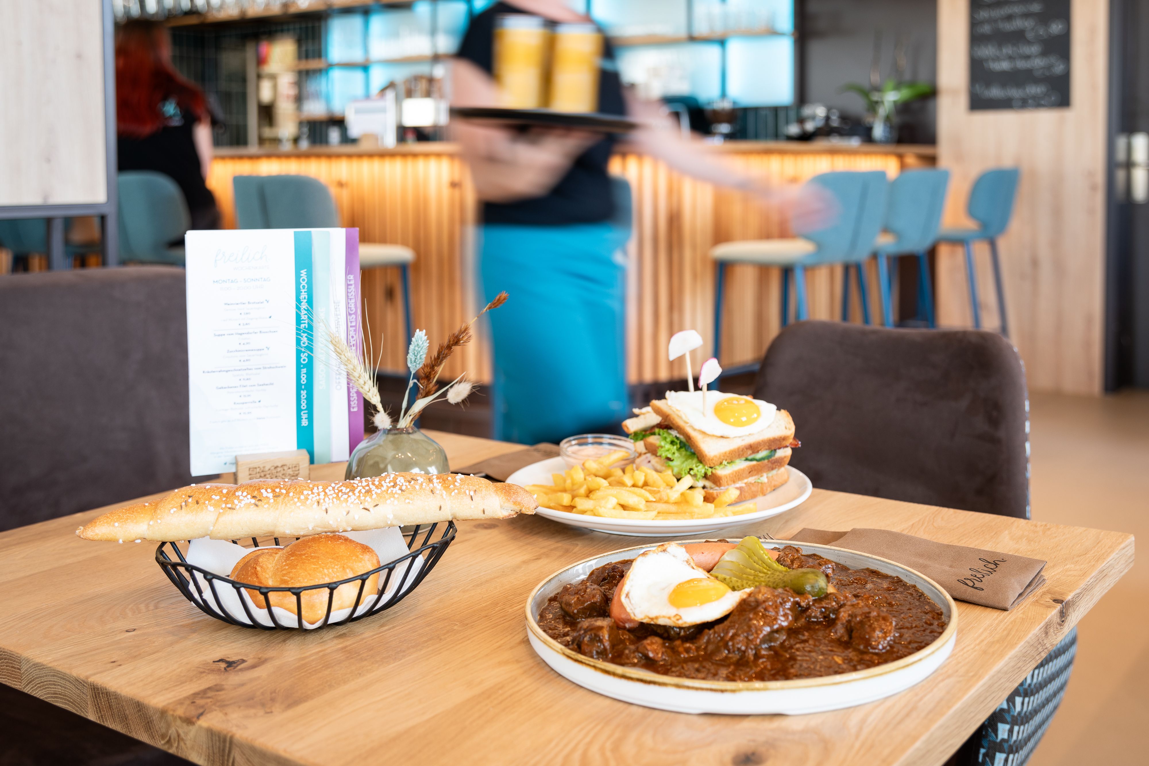 Stůl v restauraci s regionálními pokrmy: talíř guláše se smaženým vejcem, sendvič s hranolky a chlebový koš.