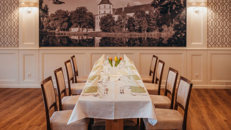 Elegantní jídelna s dlouhým stolem prostřeným bílými ubrusy a zelenými ubrousky, s velkým obrazem hradu v pozadí.