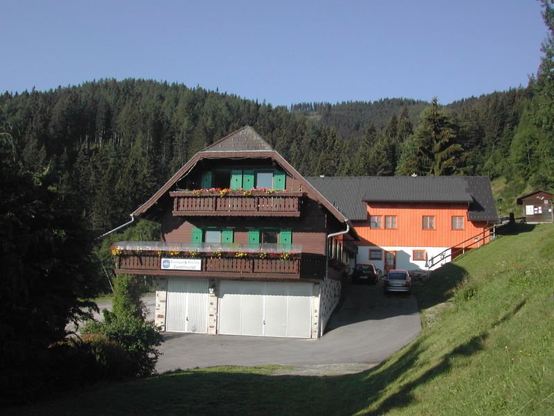 Tradiční hostinec s dřevěnými balkony a zelenými okenicemi před zalesněným kopcem.