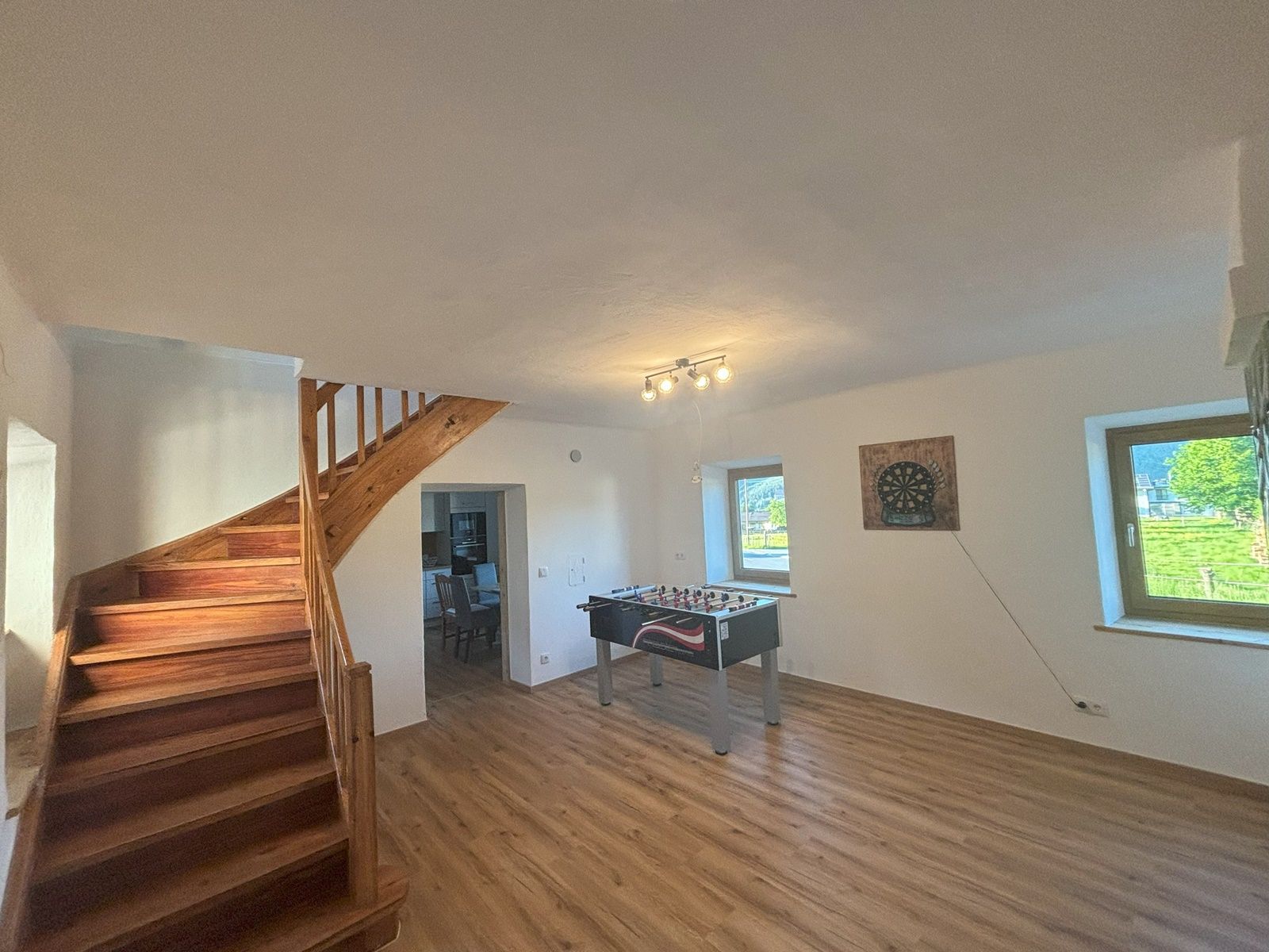 Společenská místnost se stolním fotbalem, šipkami na stěně, dřevěným schodištěm a dvěma okny.